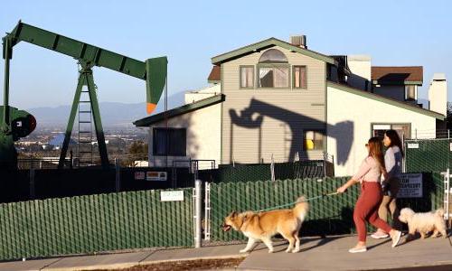 Two people and their dog walking past a house. A pumpjack is in the background.